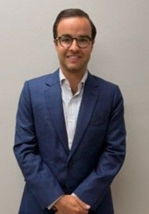 Man in a blue suit and glasses standing against a plain wall, smiling with hands clasped.