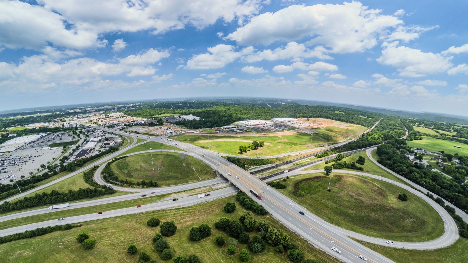 Aerial view of a highway interchange crossing green fields under a cloudy blue sky