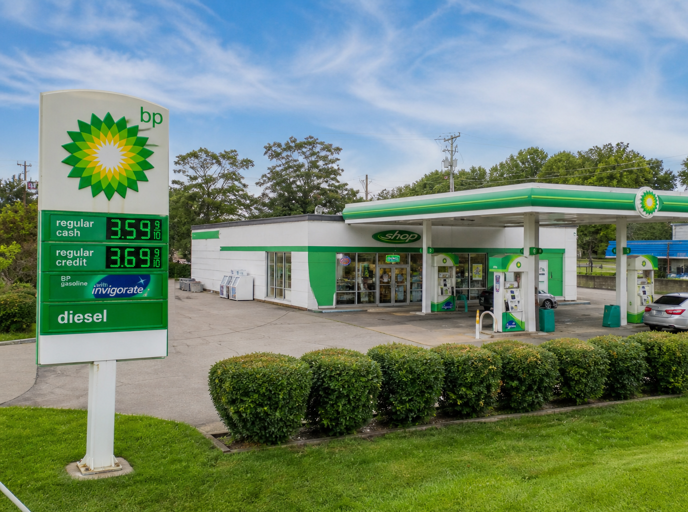 Aerial view of a gas station with a rusted red roof and parked vehicle, bordered by trees and road.
