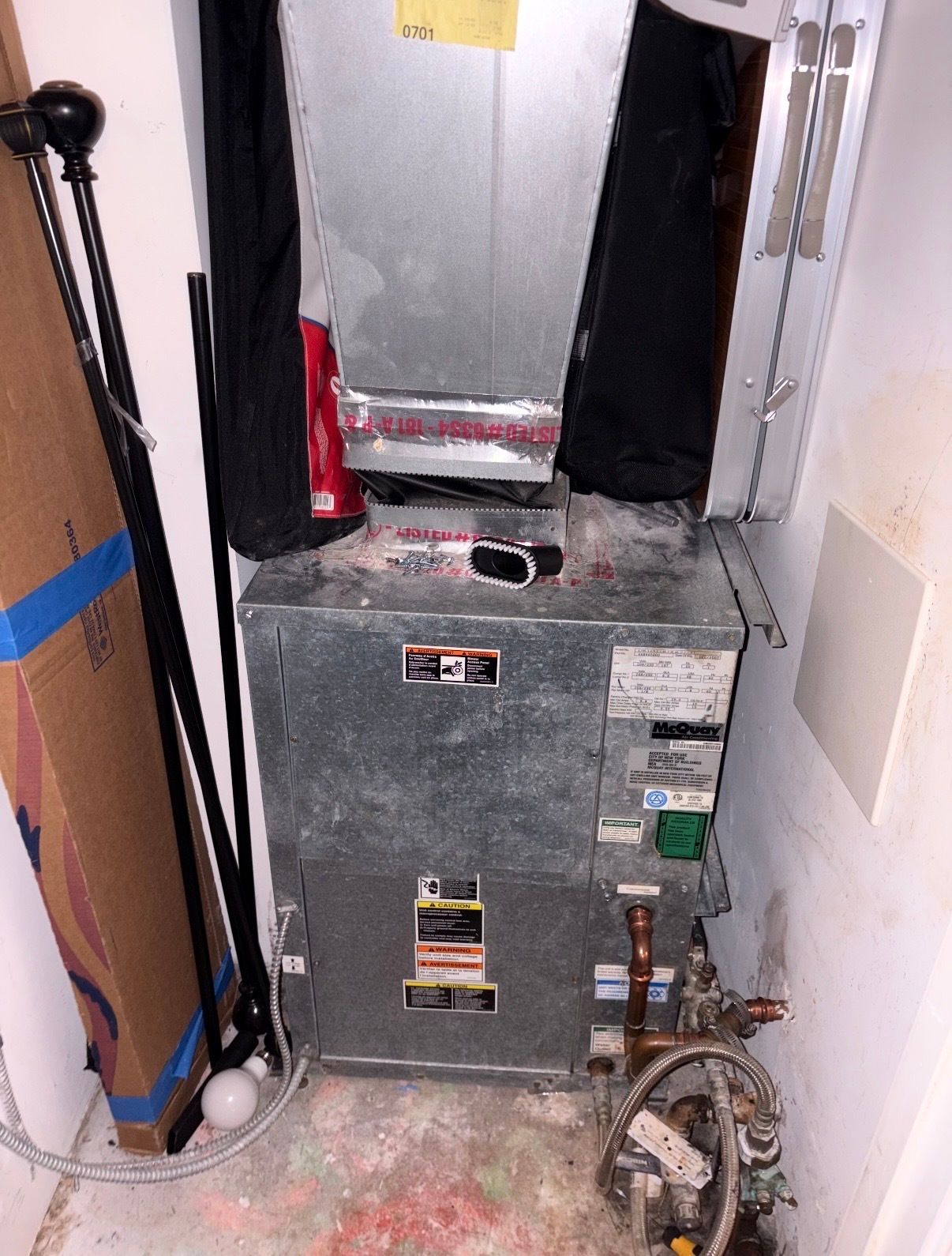 Gray HVAC furnace in a utility closet with exposed pipes and a fire extinguisher nearby.