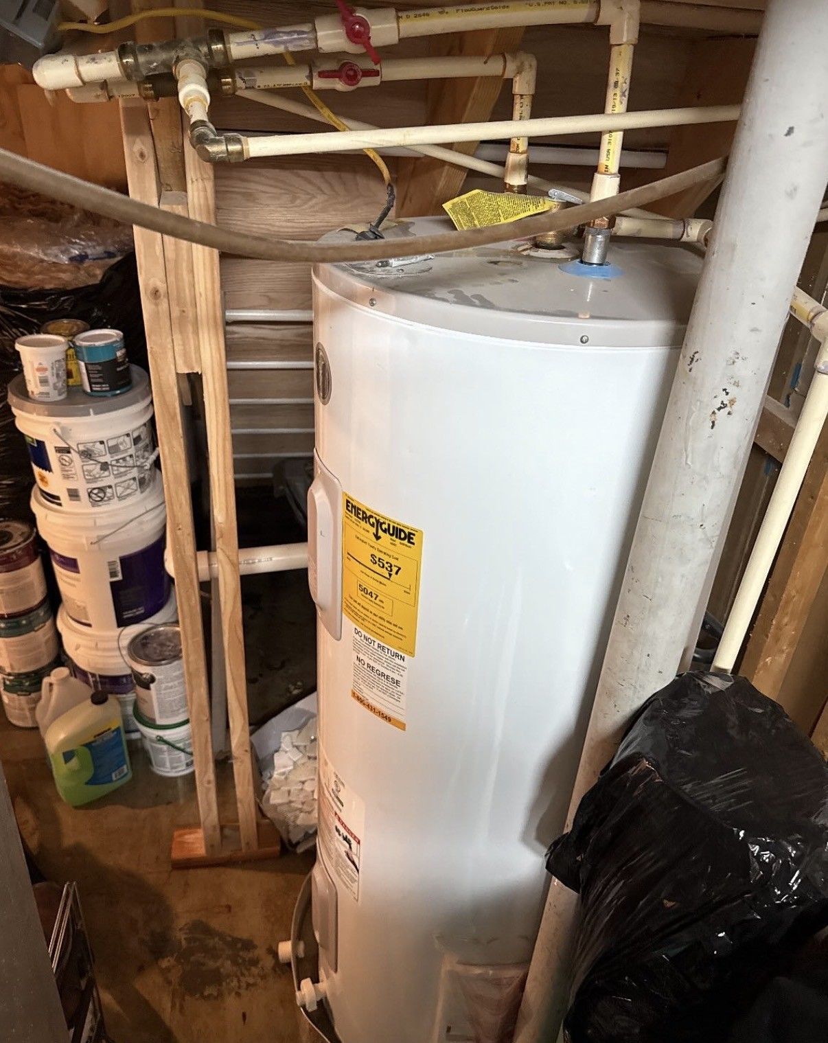 White water heater in a cluttered utility closet with pipes, stacked cans, and a black trash bag