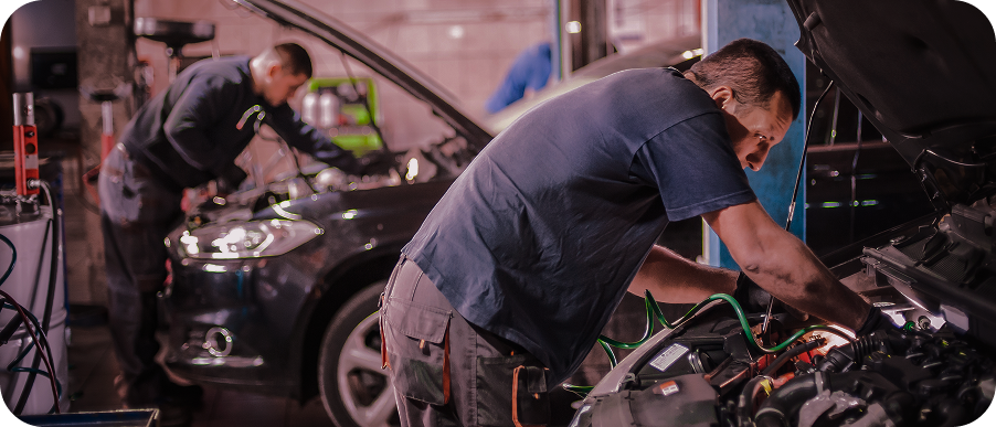 Two mechanics working under car hoods in a dim auto repair shop at night | Apex Auto Care