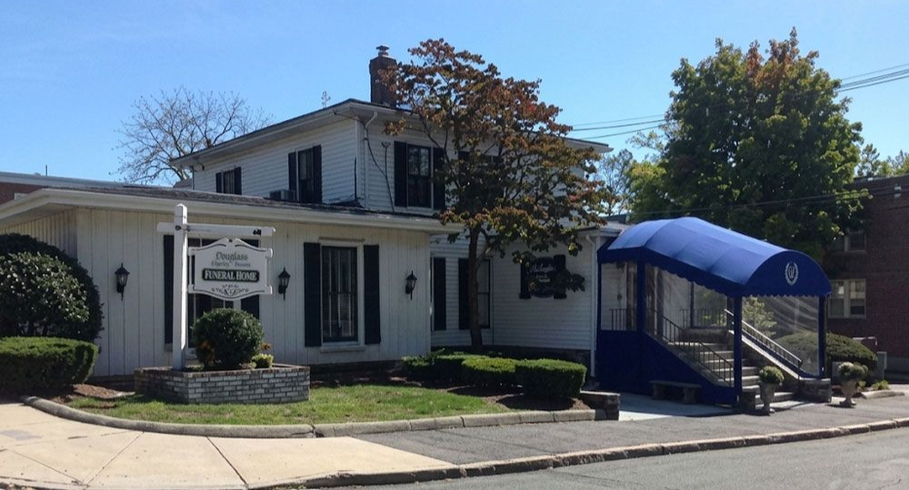 White house with black shutters and a blue awning over the front steps on a sunny street corner.
