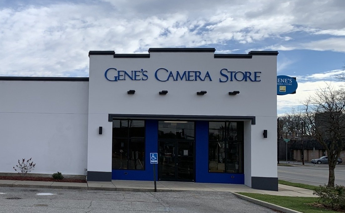 A white, single-story Gene's Camera Store building with a blue trim and entrance under a cloudy sky.