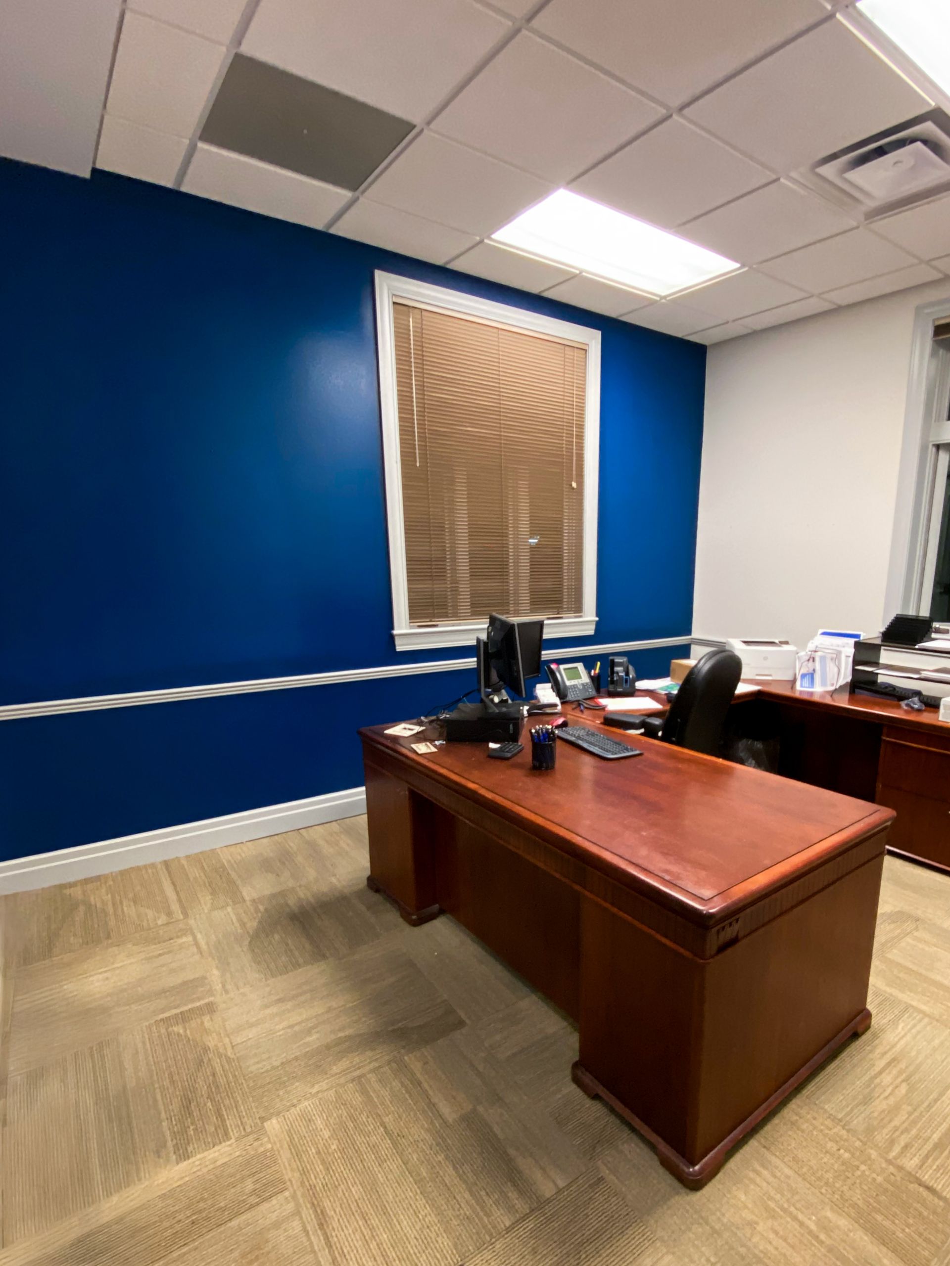 A wooden desk sits in a professional office with a deep blue accent wall, a window with blinds, and beige carpet.