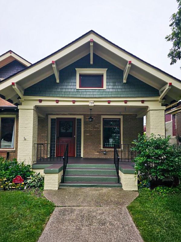 A tan bungalow with a dark green gabled roof, a front porch with stone pillars, a red door, and a concrete walkway.