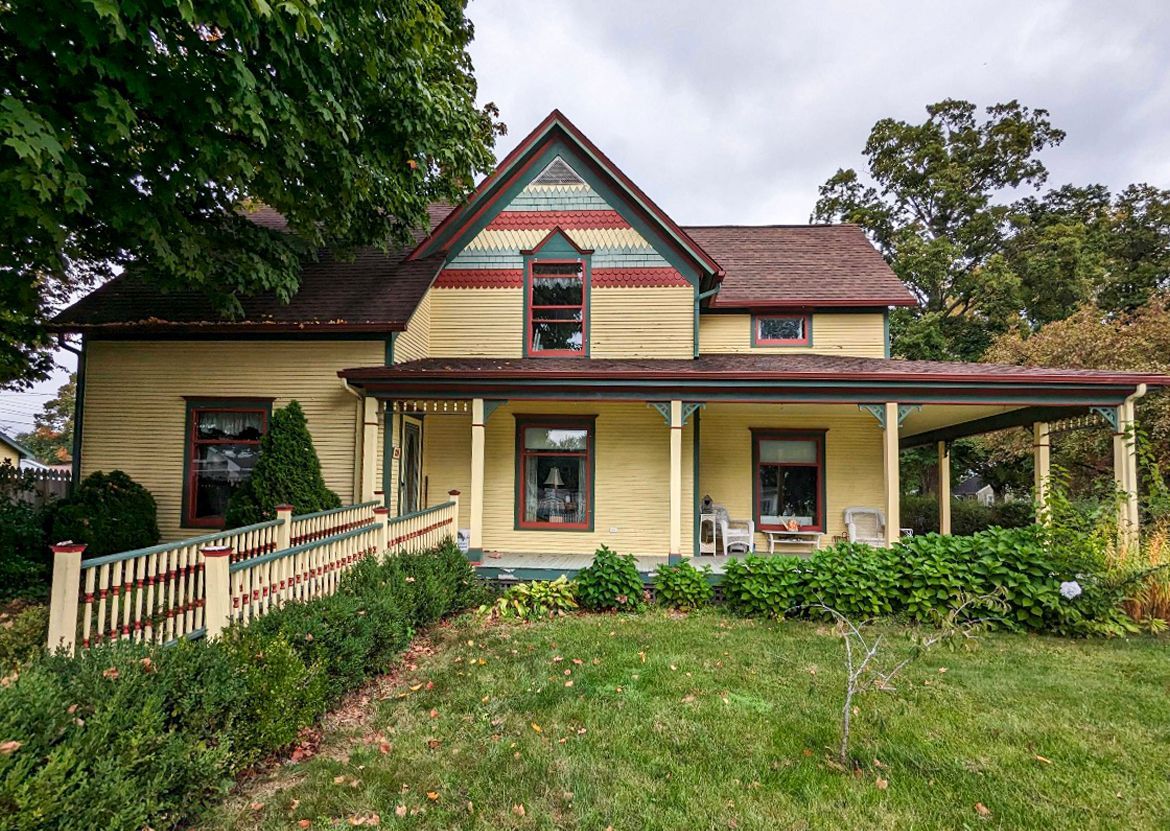 A yellow two-story house with a wrap-around porch and a wheelchair ramp, set amidst green trees and a grassy lawn.