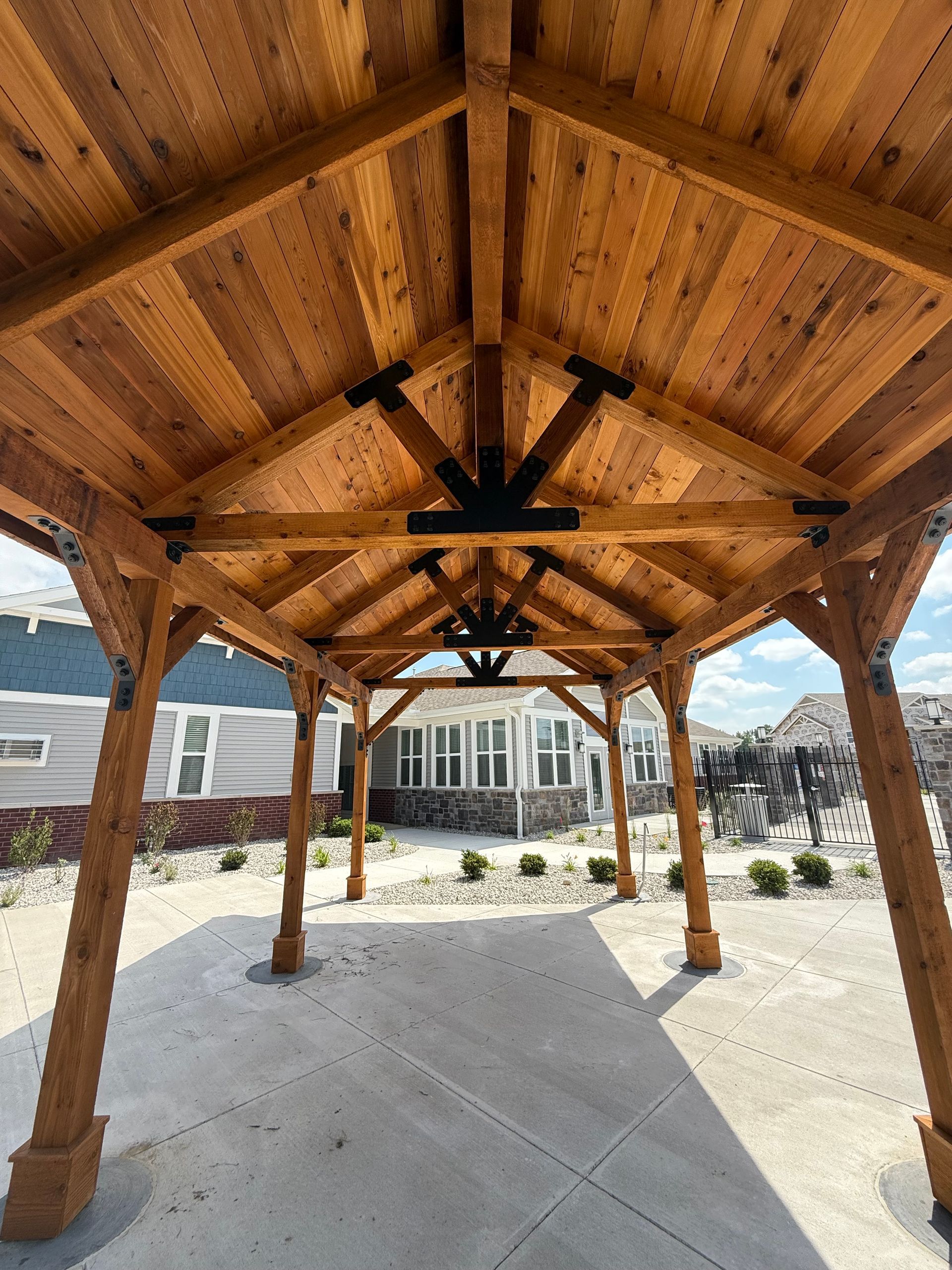 A wooden pavilion frame with exposed rafters and black metal hardware, standing over a concrete patio outside a house.