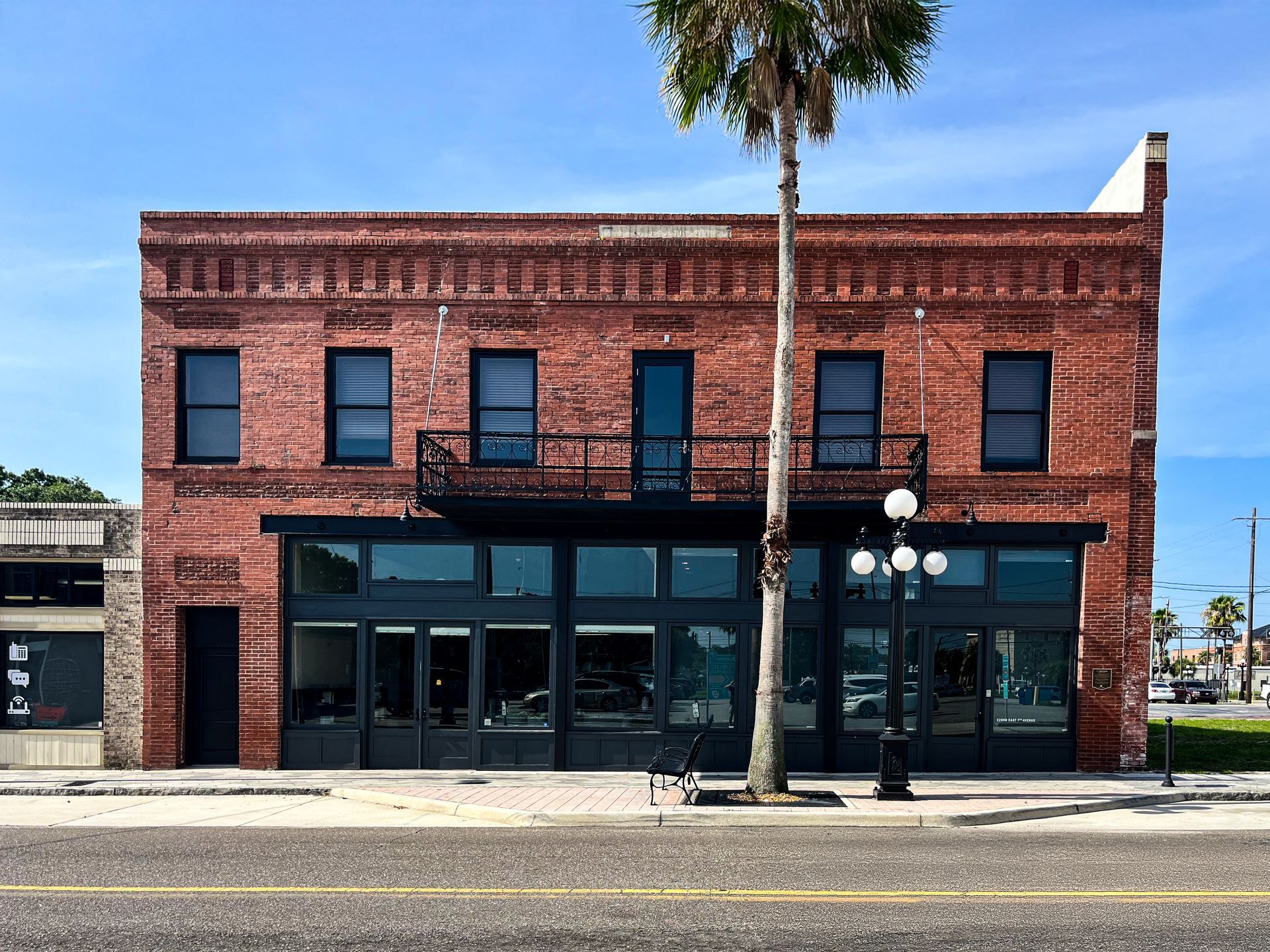 A two-story red brick commercial building with a black ground-floor storefront, a balcony, and a palm tree in front.