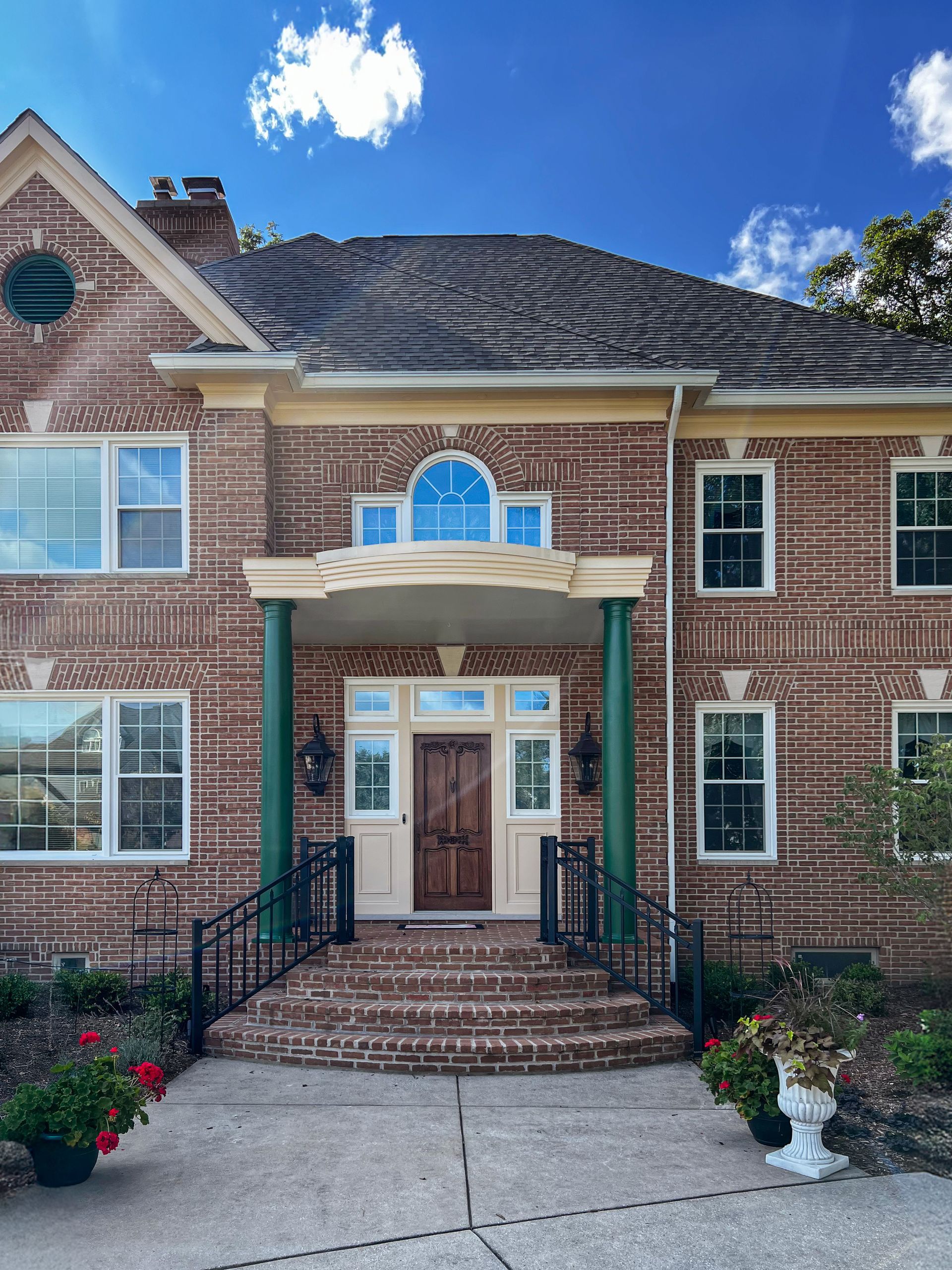 A two-story brick home with a covered front porch featuring two green columns and a wooden front door under a clear sky.