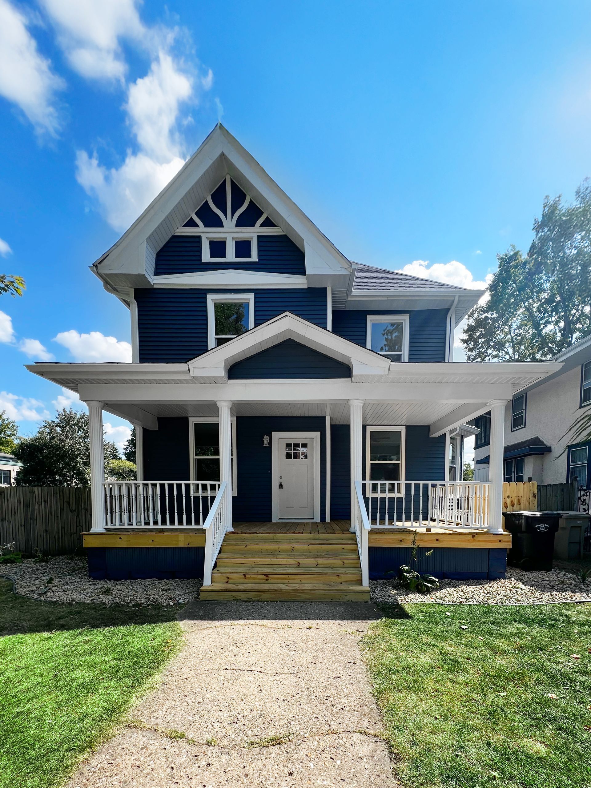 A two-story, dark blue house with white trim, a covered front porch, and wooden stairs, set under a sunny blue sky.