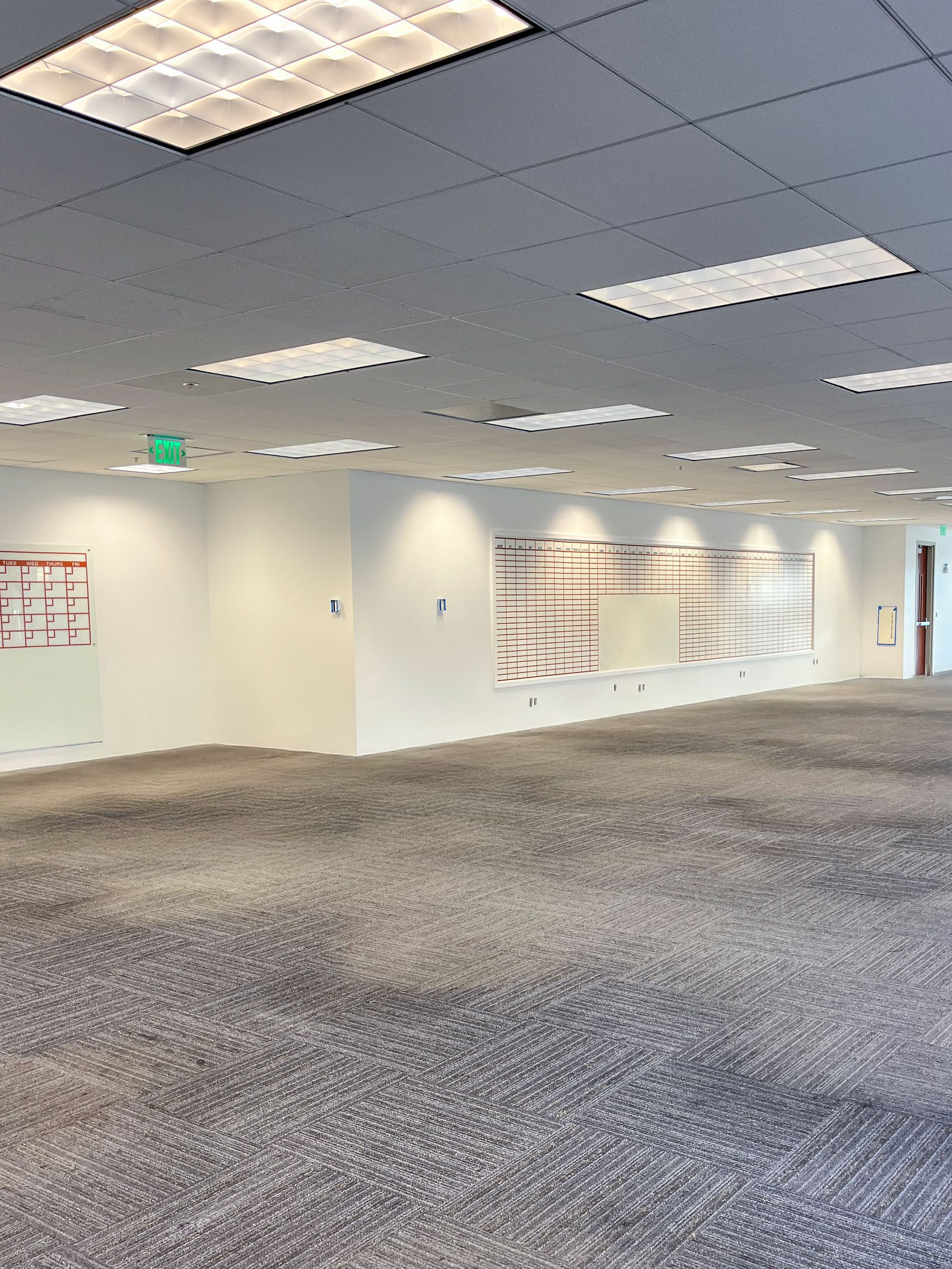 An empty office space with gray carpet, white walls, and rectangular recessed ceiling lights.