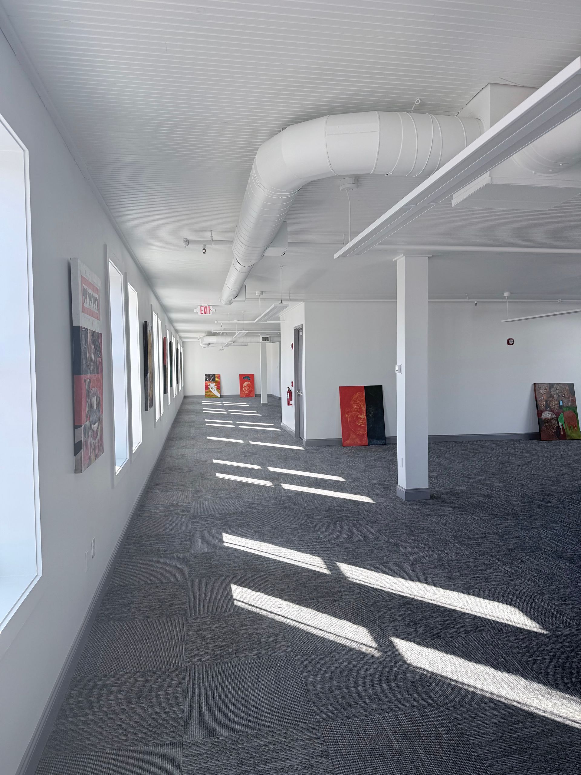 A long, sunlit hallway in an office or gallery space with grey patterned carpet, white walls, and an exposed ceiling pipe.