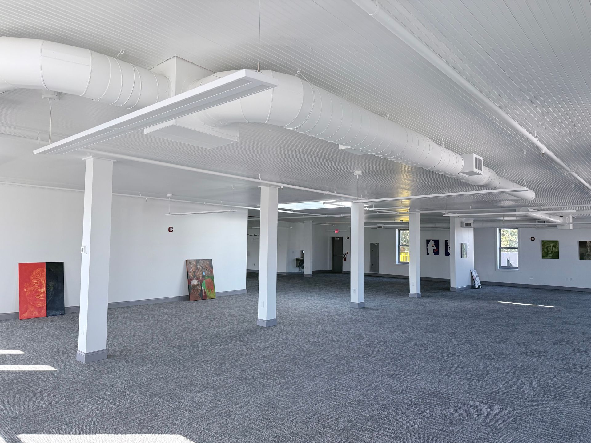 Wide-angle view of a minimalist art gallery with gray carpeted floors, white walls, support pillars, and exposed ductwork.