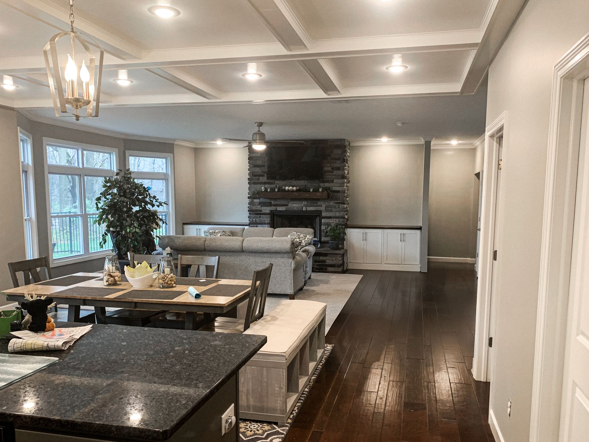 A modern open-plan living and dining area featuring dark wood floors, a stone fireplace, and coffered ceilings.