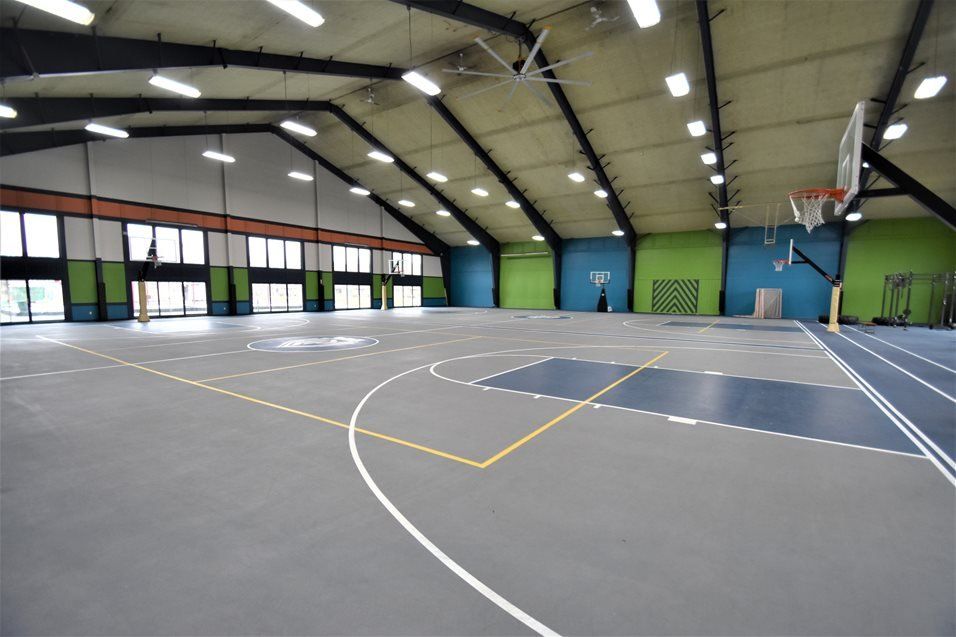 An indoor basketball court with grey flooring, yellow lines, basketball hoops, and light green and blue accent walls.