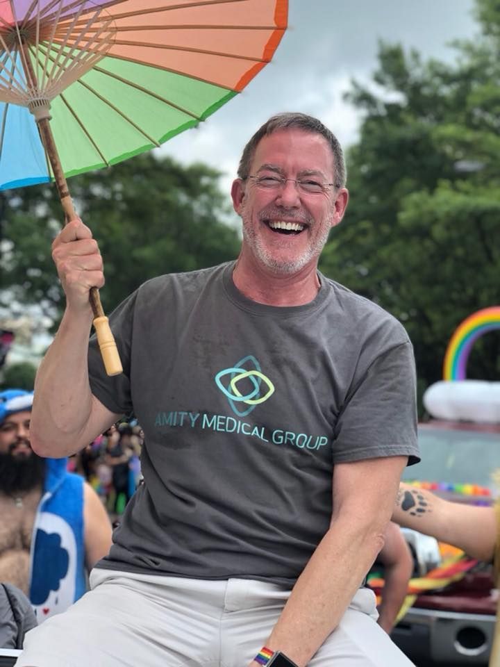 Man with gray shirt and rainbow parasol smiles at pride parade.