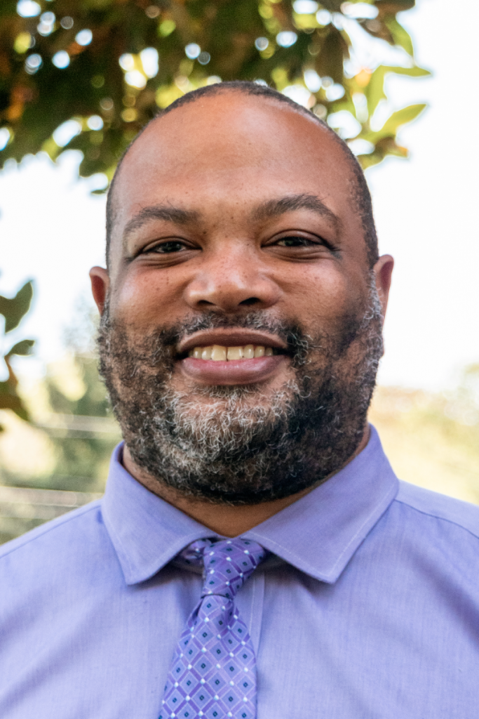 A man with a beard wearing a purple shirt and tie is smiling for the camera.