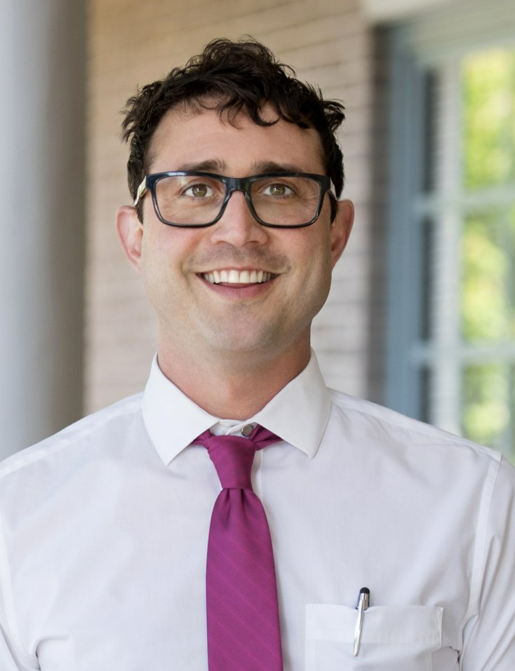 A man wearing glasses and a purple tie is smiling for the camera.