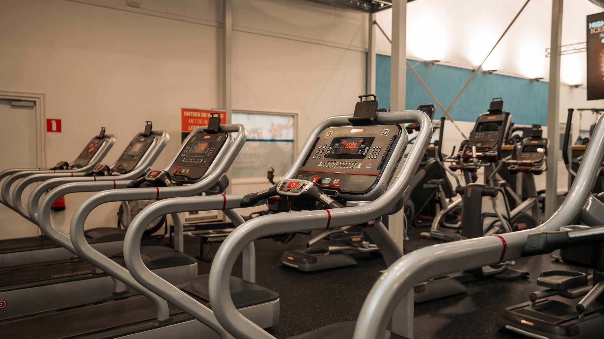 Een rij stepapparaten in een fitnesscentrum, met grijze handgrepen en zwarte schermen, en een goed verlicht interieur.