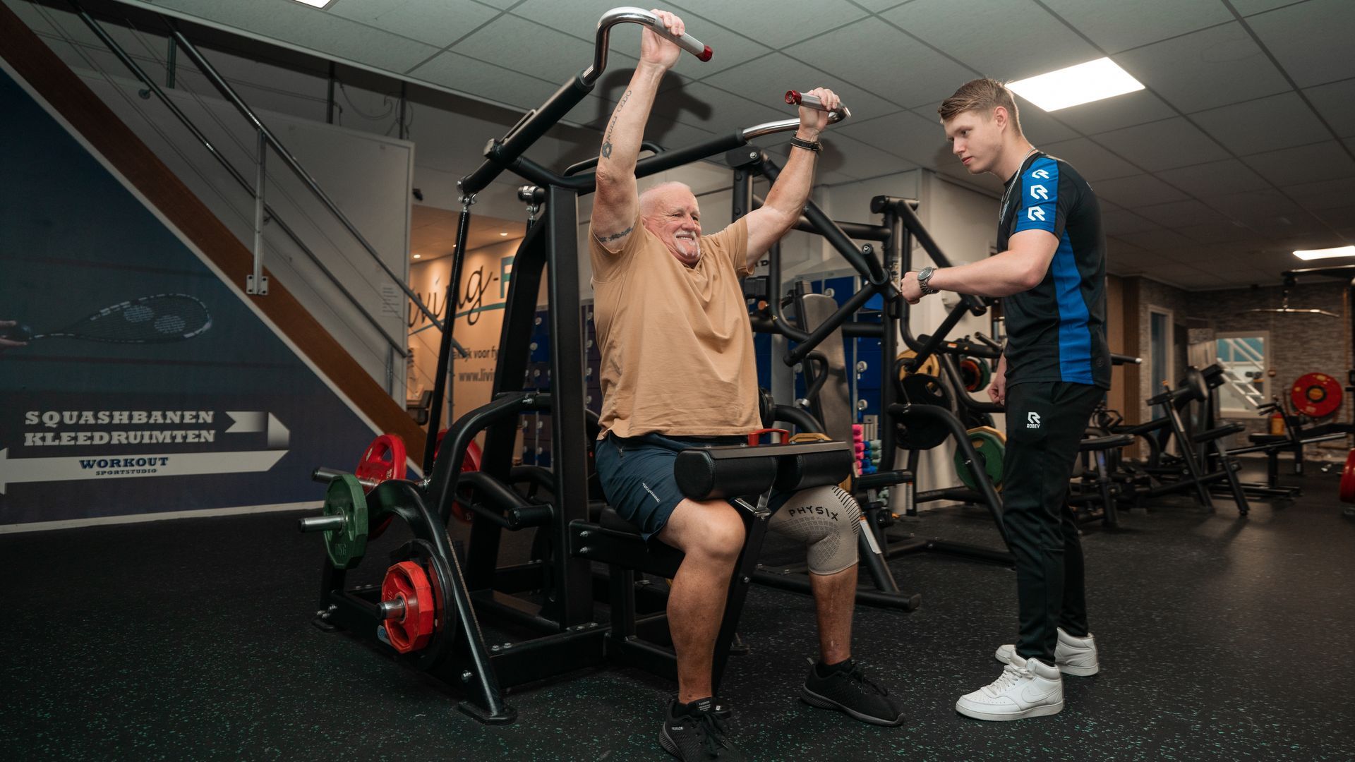Een oudere man traint met een trainer op een apparaat in de sportschool.