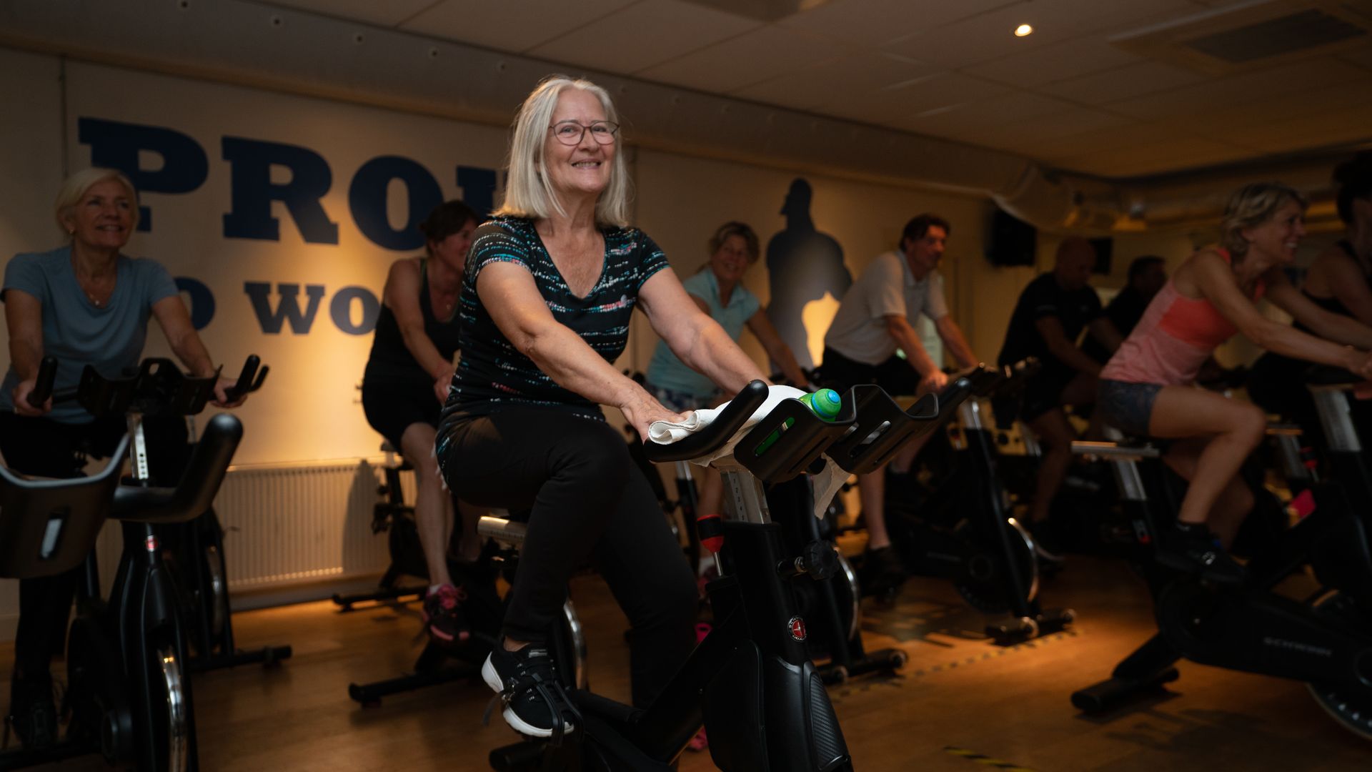 Mensen fietsen in een spinningles. De vrouw vooraan glimlacht; anderen sporten in een schemerig verlichte ruimte.