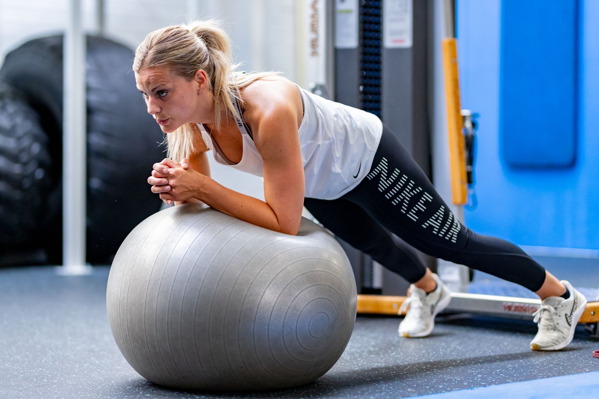 Een vrouw doet een plank op een fitnessbal in de sportschool. Ze draagt ​​Nike-kleding en ziet er geconcentreerd uit.