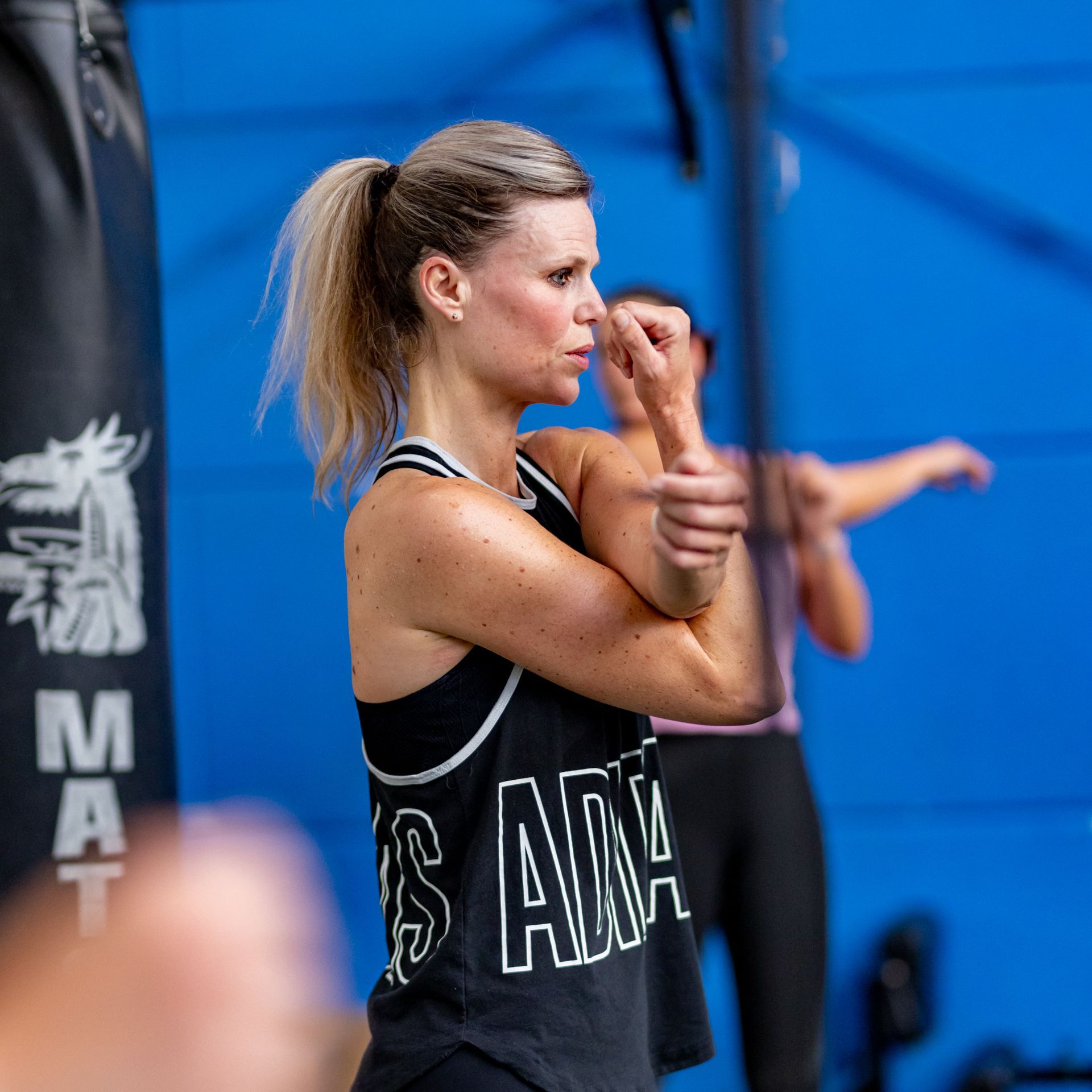 Een vrouw in de sportschool strekt haar arm uit; ze draagt ​​een zwart tanktopje met witte letters.