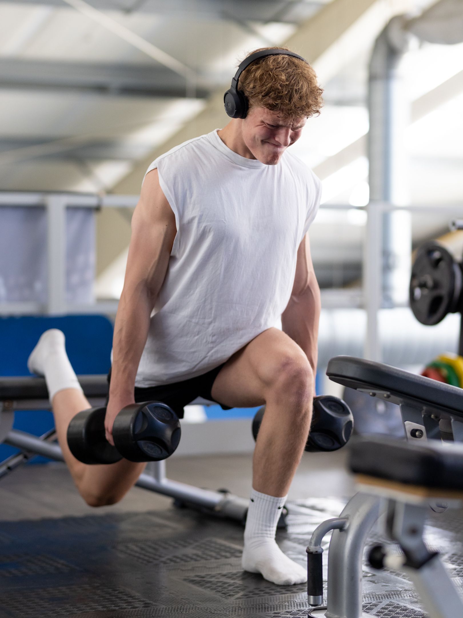 Man doet dumbbell lunges in de sportschool, draagt ​​een koptelefoon, een wit shirt, sokken en een zwarte korte broek.