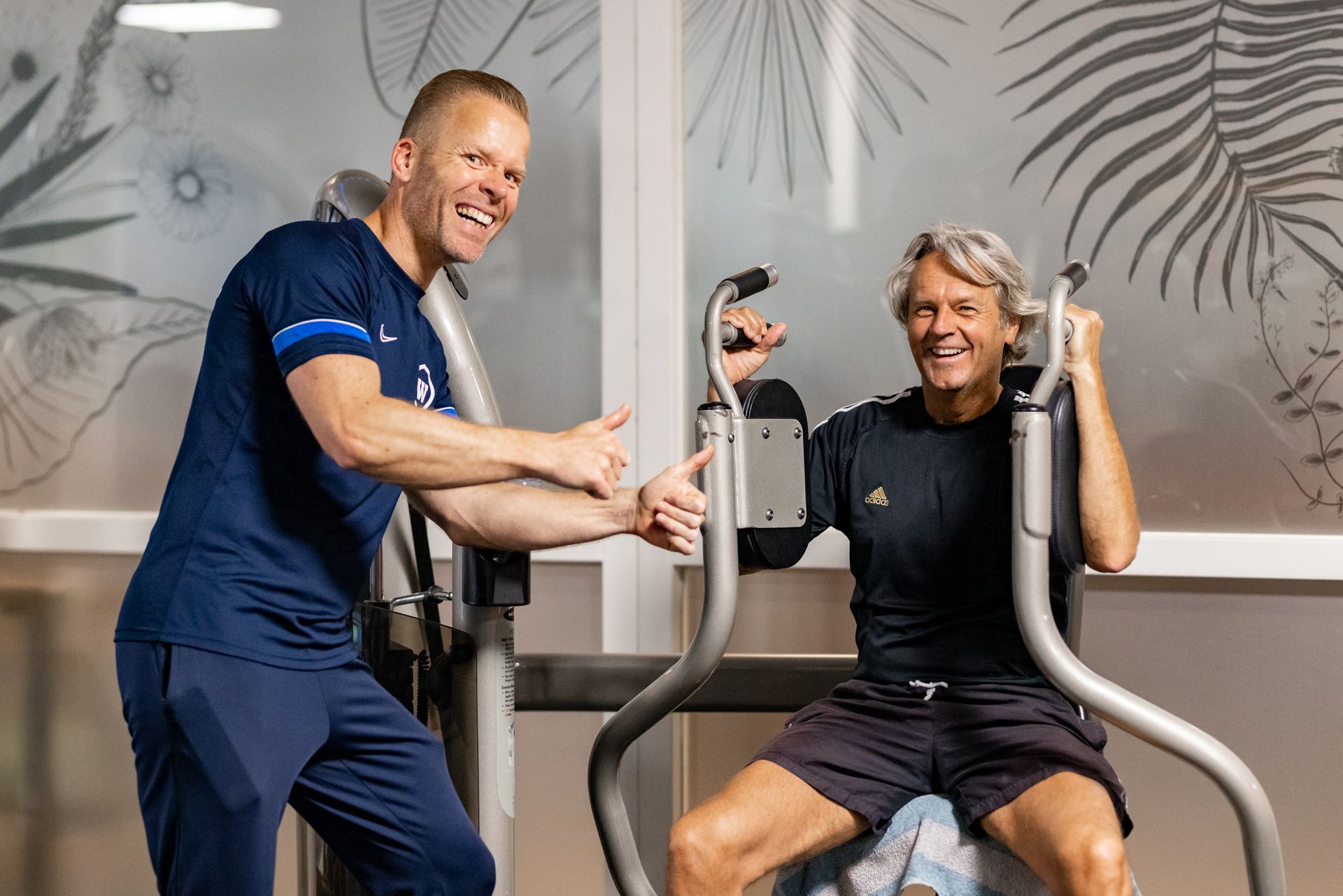 Man traint een andere man op een fitnessapparaat. Ze lachen allebei en steken hun duimen omhoog in de sportschool.