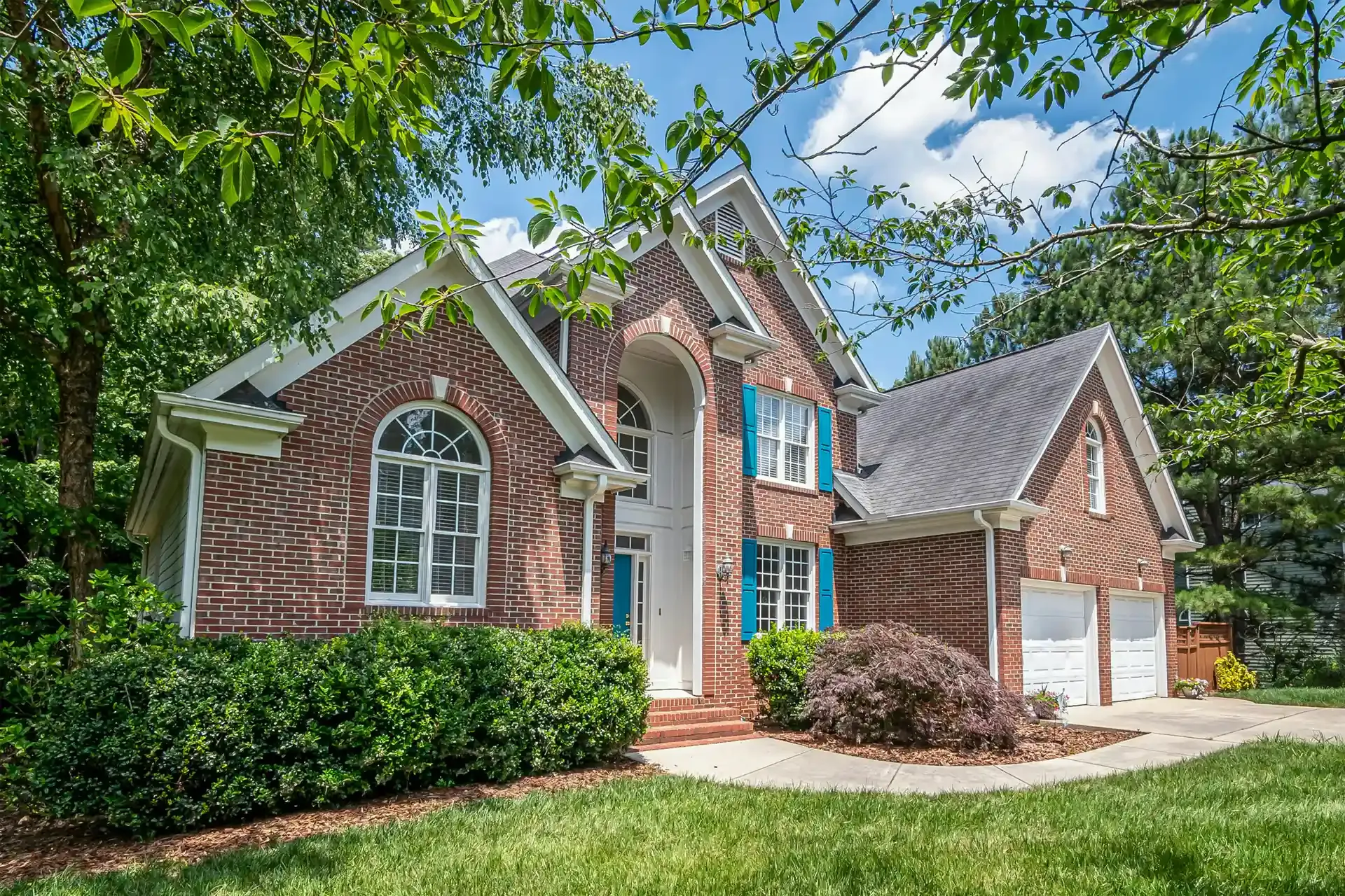 Brick suburban house with white trim, arched windows, and a two-car garage surrounded by green trees and lawn