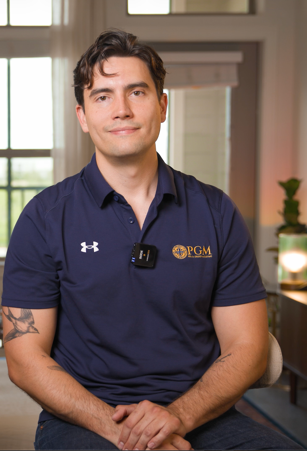 Man seated indoors in a navy polo shirt with a lapel mic, facing the camera