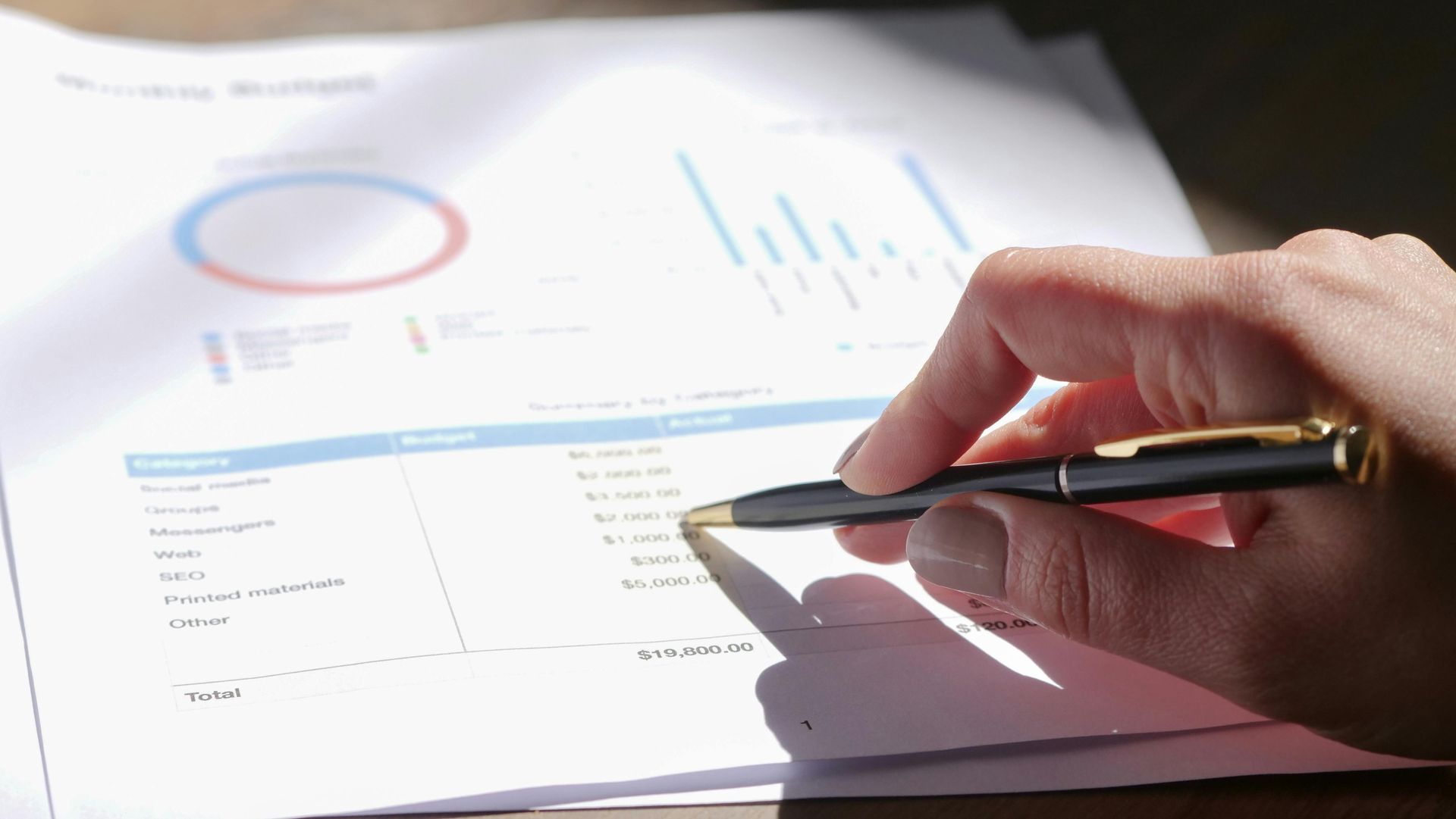 Hand holding a pen points at financial data on a printed document with charts.