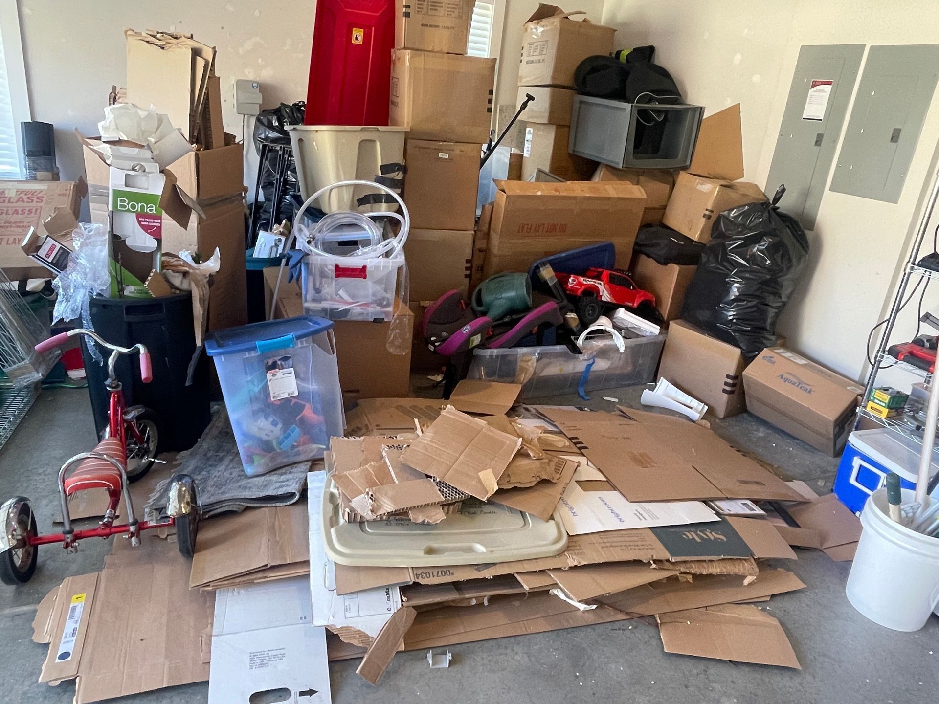 Cluttered garage or storage room filled with stacked boxes, bags, cardboard, and scattered household items