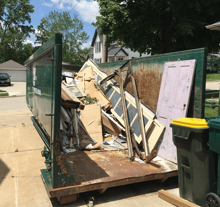 Open trailer loaded with broken drywall and wood debris beside a green trash bin on a driveway