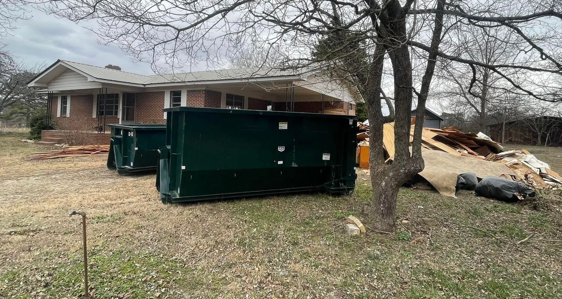 Large green dumpster in a yard beside a damaged house with fallen debris and bare trees