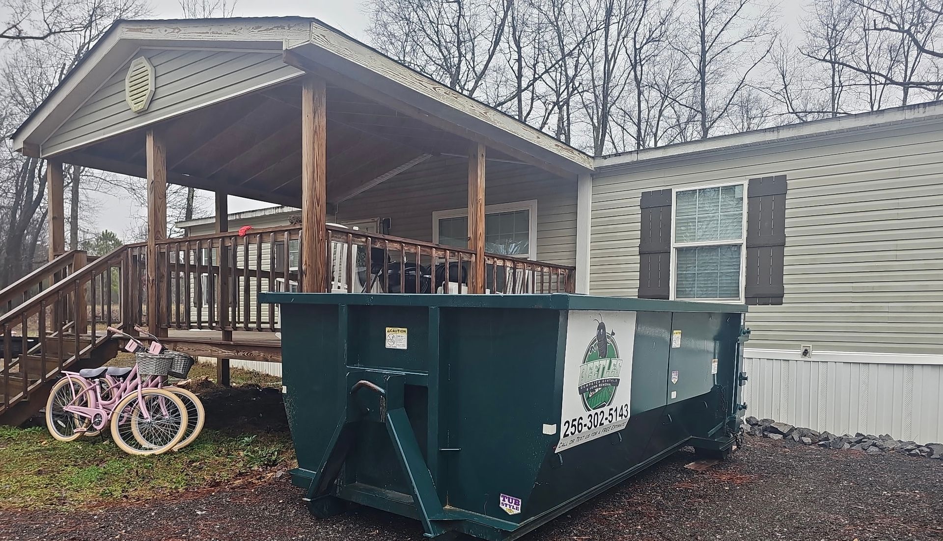 Green dumpster beside a porch with bicycles, in front of a house on a cloudy day