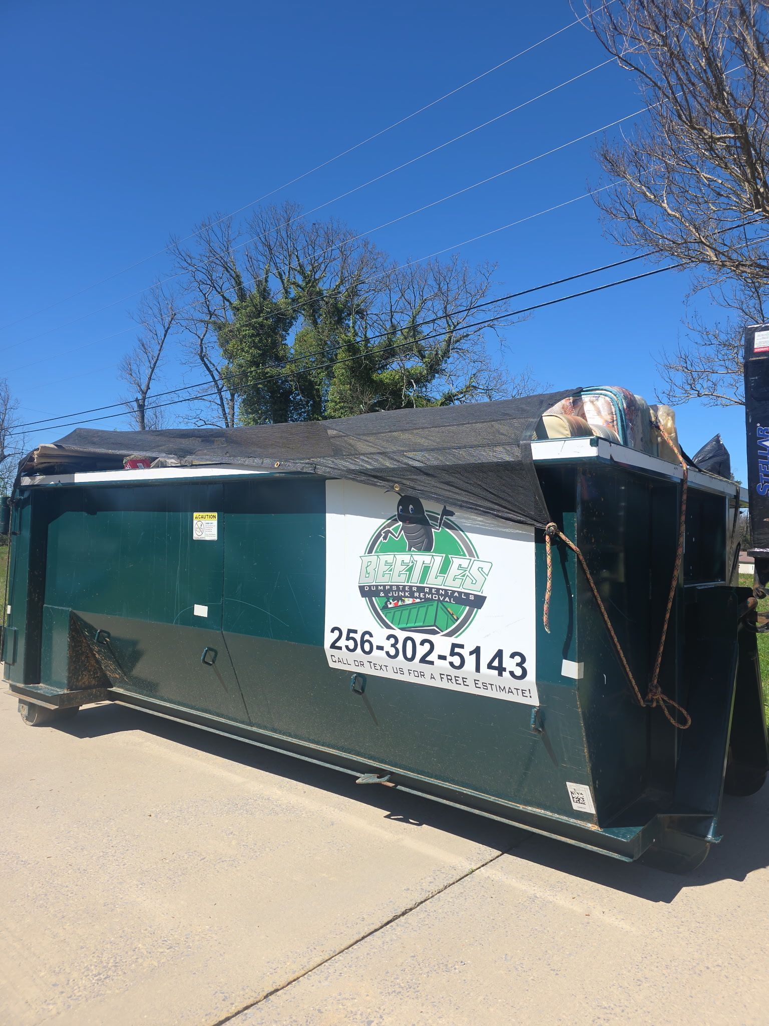 Green dumpster on a driveway under a blue sky, with a white company sign and phone number.