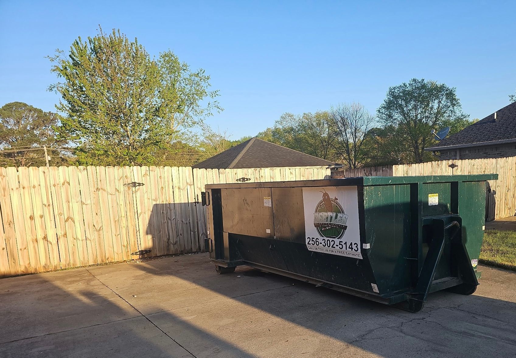 Green dumpster in a fenced parking lot under a clear blue sky, with a small shed nearby.