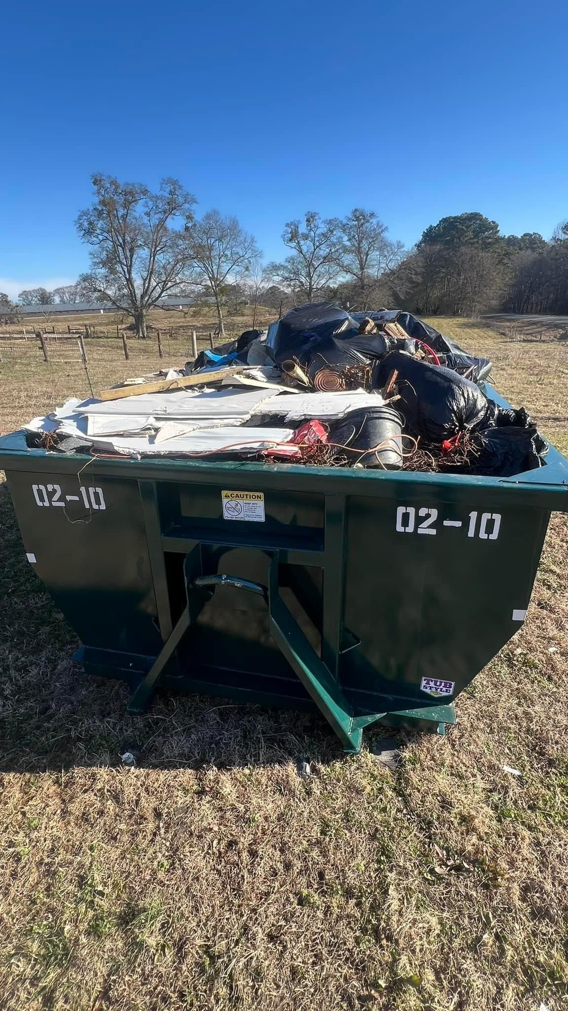 Green dumpster filled with debris in a dry, grassy field under a blue sky