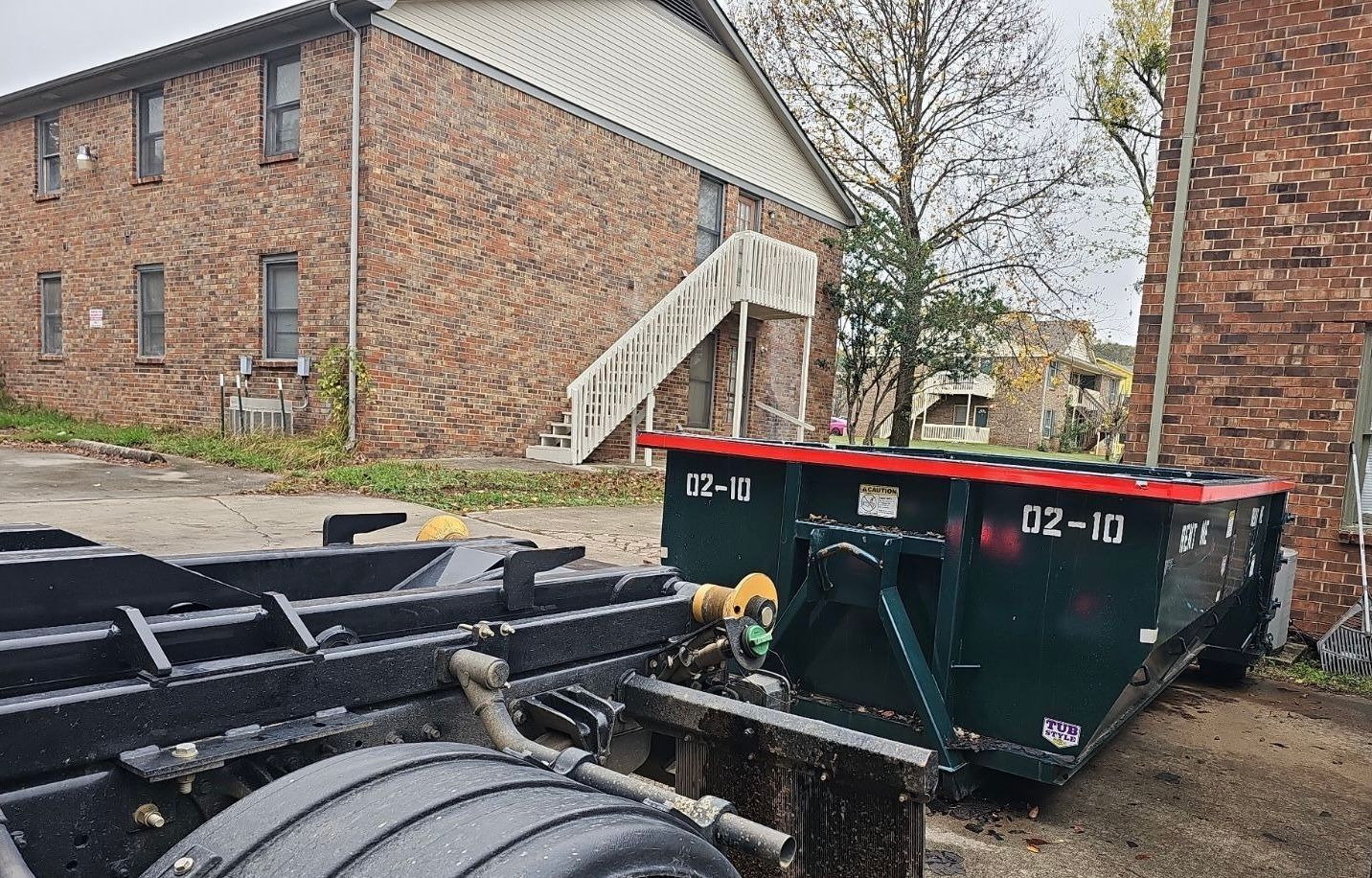 Large black dumpster beside a brick apartment building with a white stairway on a wet day