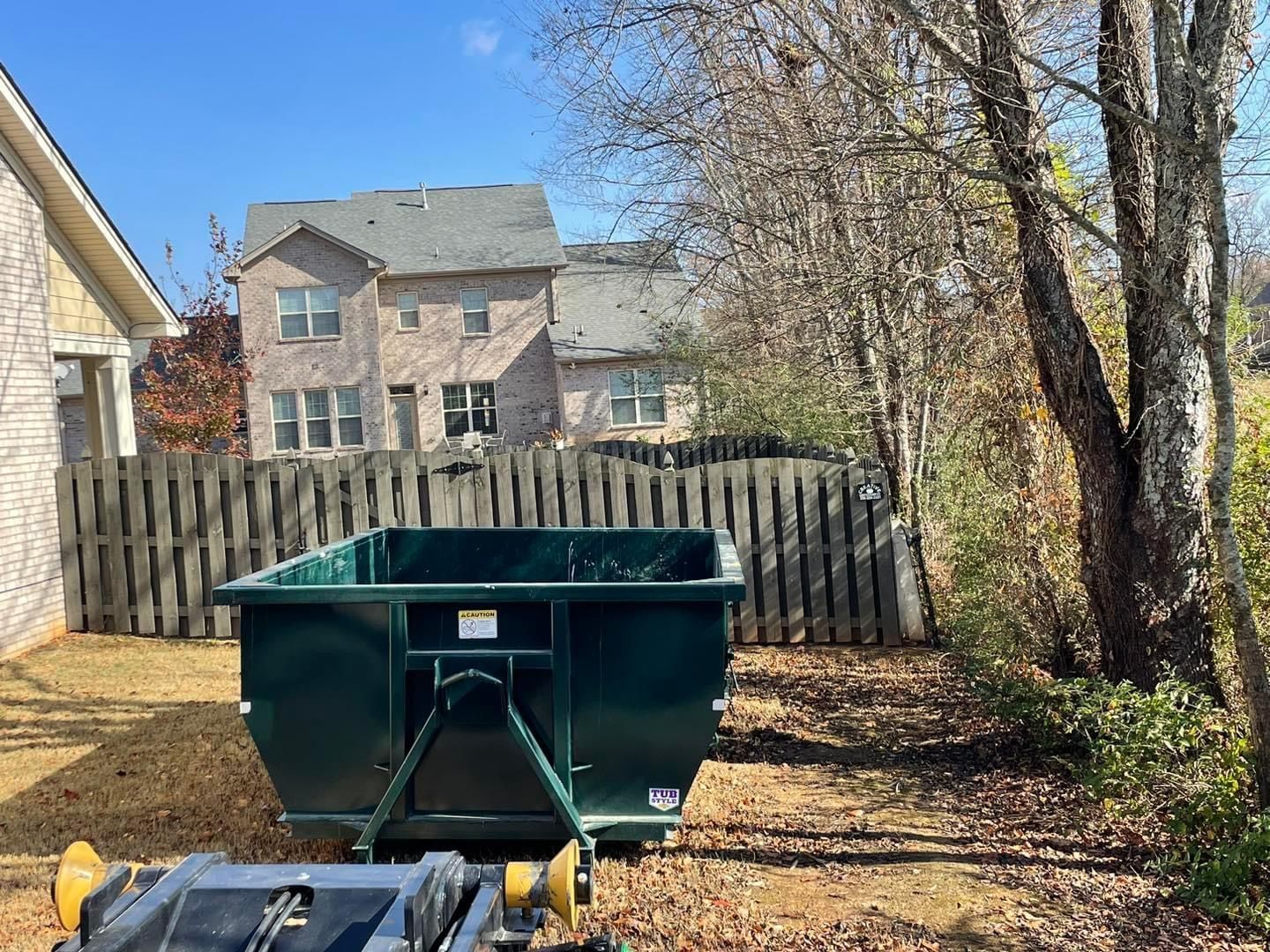 Green dumpster in a fenced backyard with a house and trees in the background