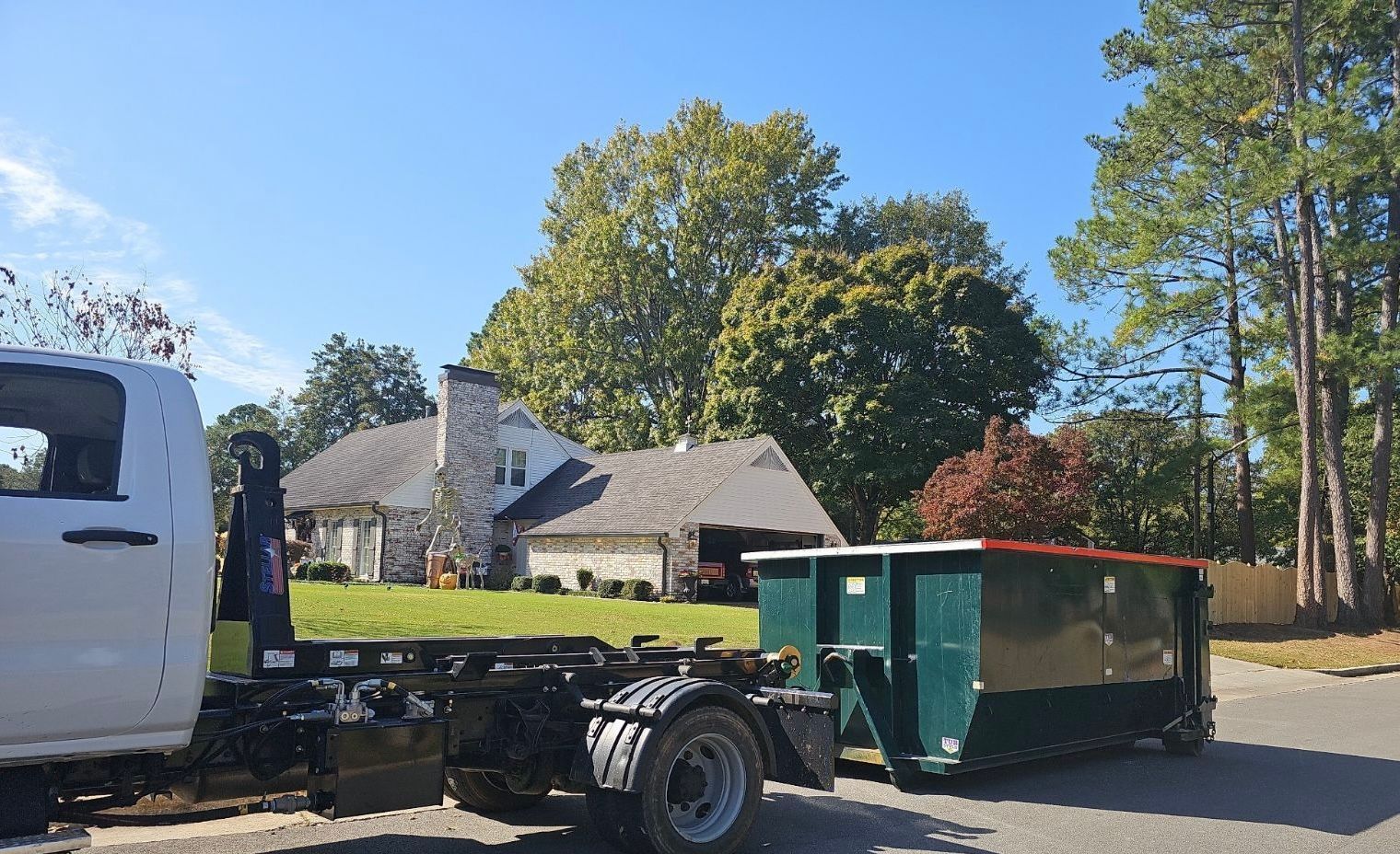 Construction dump trailer beside a flatbed truck in a sunny suburban yard with a house and trees behind.