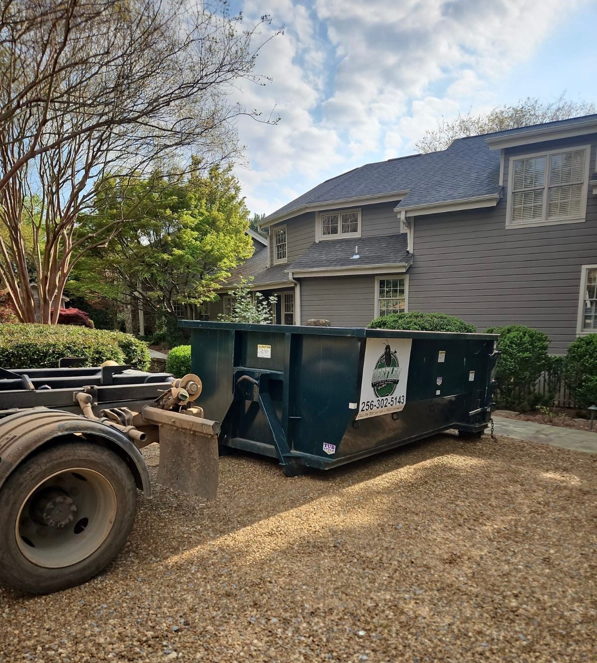 Green dumpster beside a gray house, with a truck in the foreground on a gravel driveway.