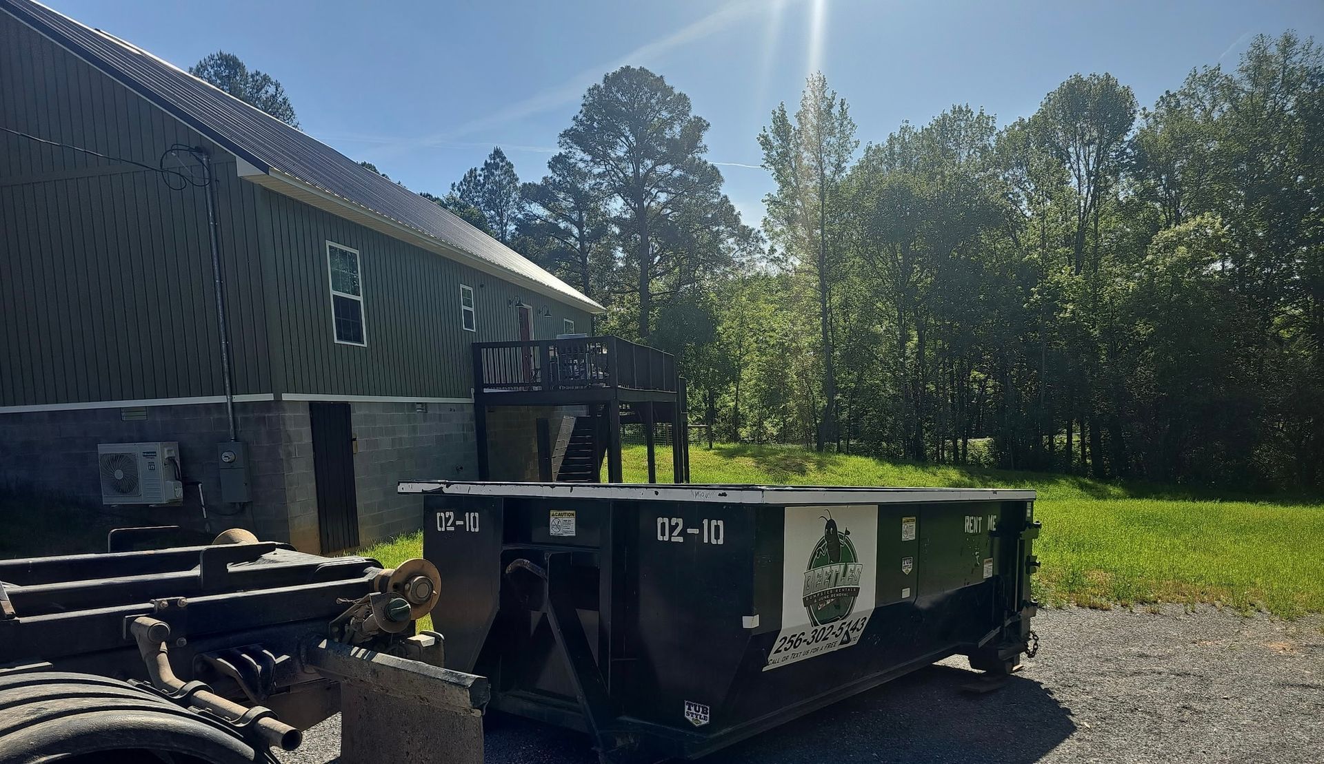 Green dumpster beside a dark building in a sunny yard with trees and grass.