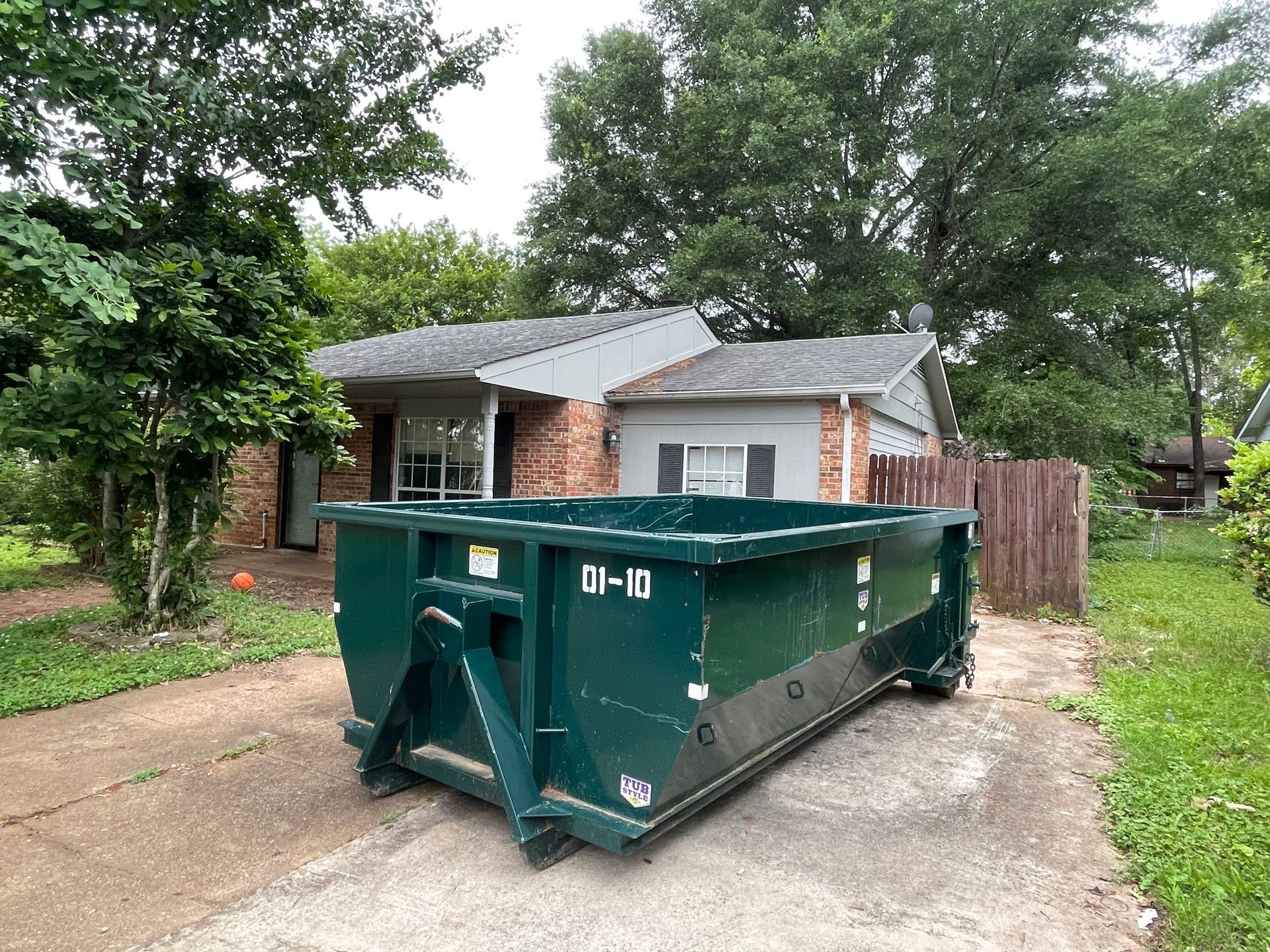 Green dumpster in a driveway in front of a house with trees and bushes.