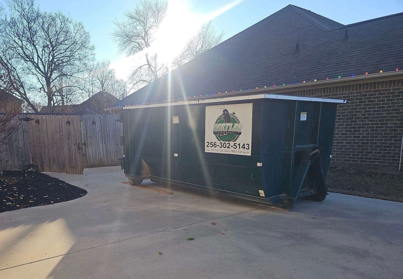 Dark green dumpster in a driveway beside a brick house, with sunlight and trees in the background.