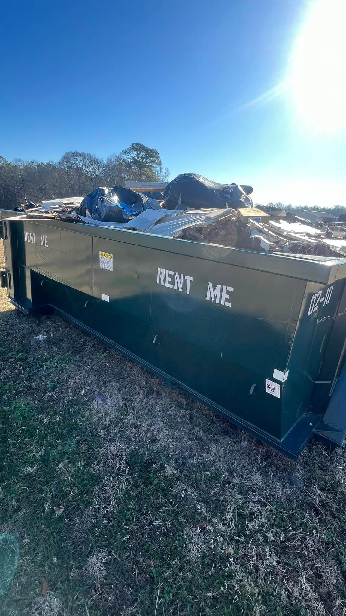 Large green dumpster labeled “DEBT MOE” in a sunny gravel lot, with rocks and trees behind it.
