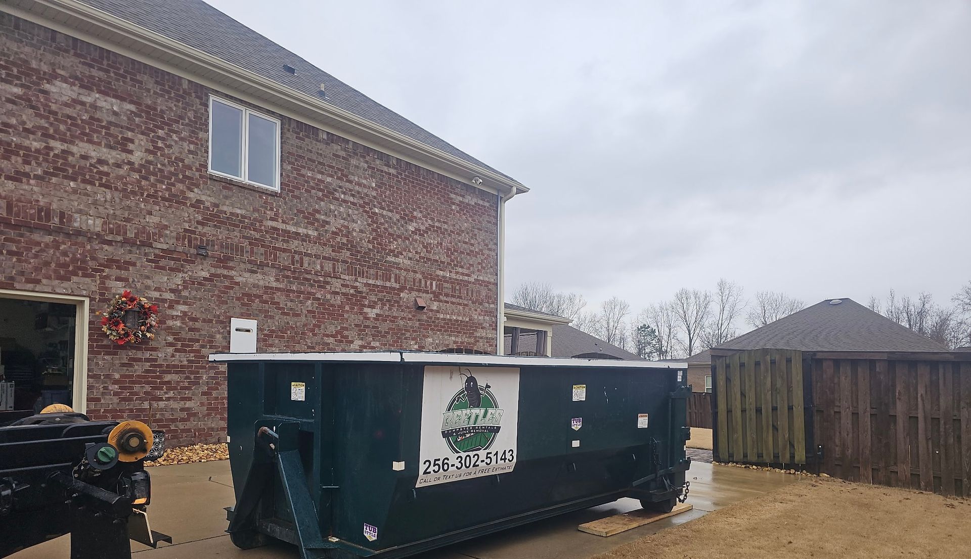Large green dumpster beside a brick house in a fenced backyard on a cloudy day