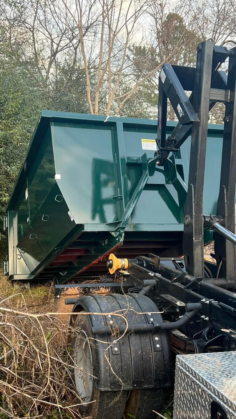 Green dump trailer tipped on its side beside heavy equipment in a wooded area
